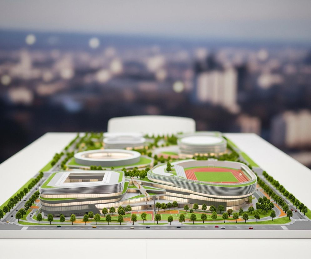Na zdjęciu centralnie umieszczony jest szczegółowy model architektoniczny kompleksu sportowo-rekreacyjnego, umieszczony na białym stole. Model przedstawia kilka budynków o nowoczesnej architekturze, z dachami pokrytymi zielenią, otoczone trawnikami, rzędami małych drzew i drogami z miniaturowymi samochodami. Po lewej stronie dominuje prostopadłościenny stadion z jasnoszarą elewacją, a po prawej owalny stadion z czerwoną bieżnią i zielonym boiskiem, również o zielonym dachu. W tle, rozmyte tło przedstawia nocną panoramę miasta z licznymi, rozświetlonymi budynkami.