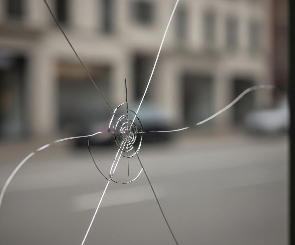 Przednie szyba ma w środku punkt uderzenia, od którego rozchodzą się promieniście pęknięcia. Spiralne pęknięcia tworzą mały okrąg w centrum, z którego wychodzą dłuższe, nieregularne linie rozciągające się na całej powierzchni szyby. Tło jest rozmyte, ukazując szare budynki z ciemnymi oknami i zamazane kształty pojazdów. Po prawej stronie kadru widoczna jest ciemna, pionowa rama okienna.