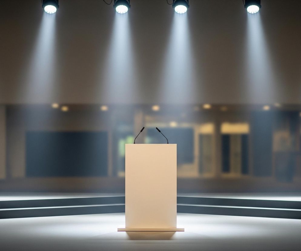 Na pierwszym planie, centralnie, stoi beżowa mównica z dwoma czarnymi mikrofonami skierowanymi ku górze. Podłoże przed mównicą jest jasnoszare i odbija światło, za nią zaś znajdują się dwa ciemne, poziome stopnie. W tle widać rozmyte, ciemne wnętrze pomieszczenia z elementami przypominającymi okna lub witryny, częściowo oświetlone od wewnątrz. Od góry, z sufitu, zwisa pięć okrągłych reflektorów, z których każdy emituje stożkową wiązkę jasnego światła w dół.