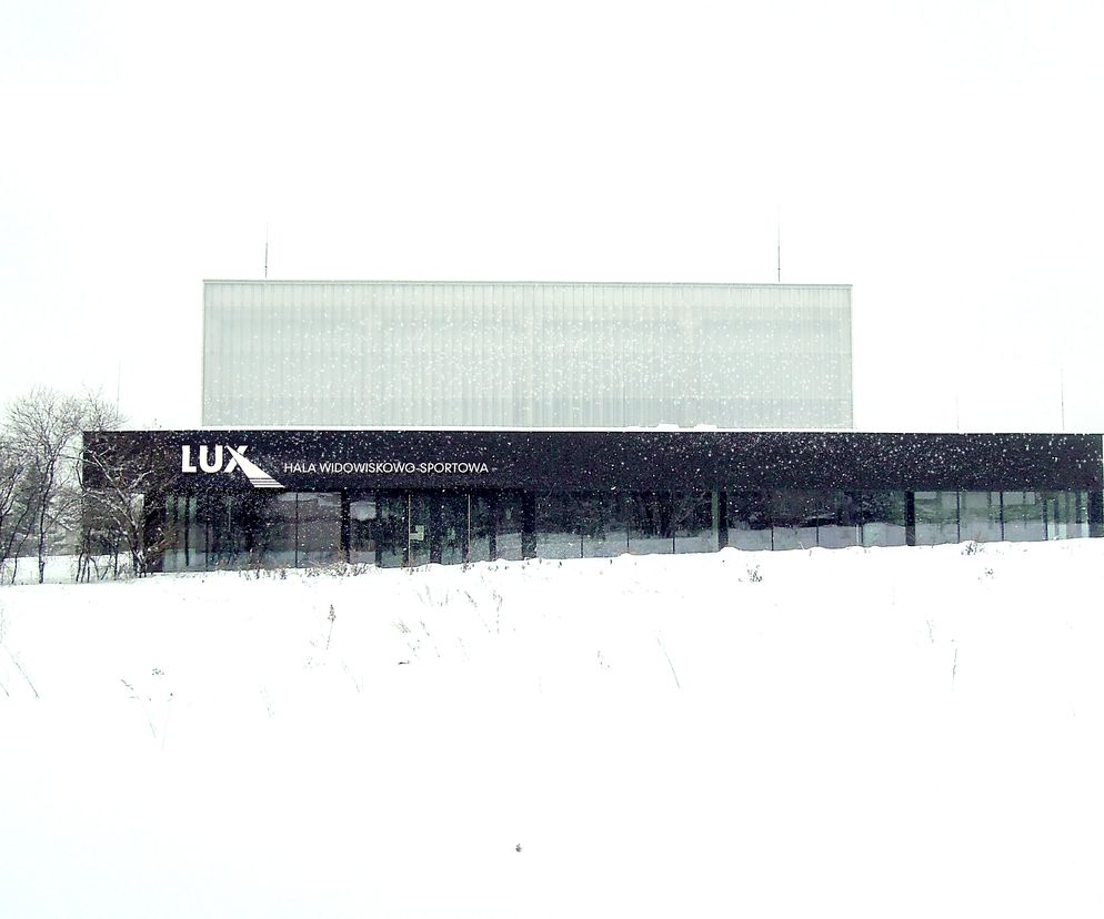 Hala widowiskowo-sportowa LUX w Rogoźniku, widziana zimą, z frontową elewacją z ciemnego szkła i transparentną częścią sportową, otoczona ośnieżonym terenem i drzewami. Więcej o projekcie na portalu Architektura Murator Plus.