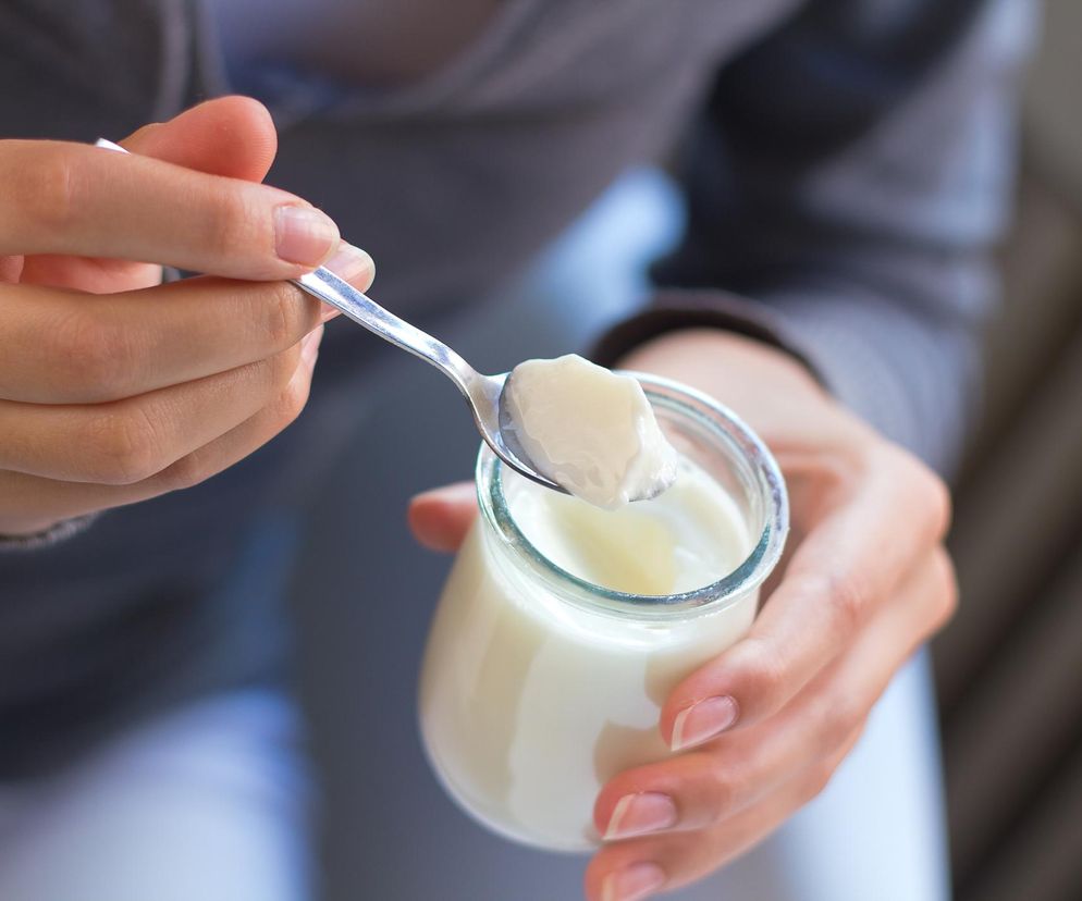 Dłonie trzymające otwarty szklany słoik z naturalnym jogurtem, z metalową łyżeczką nabierającą porcję produktu. Scena symbolizuje zdrowe odżywianie i dbałość o jelita, co ma znaczenie w kontekście profilaktyki choroby Crohna, o czym przeczytasz na Poradnik Zdrowie.