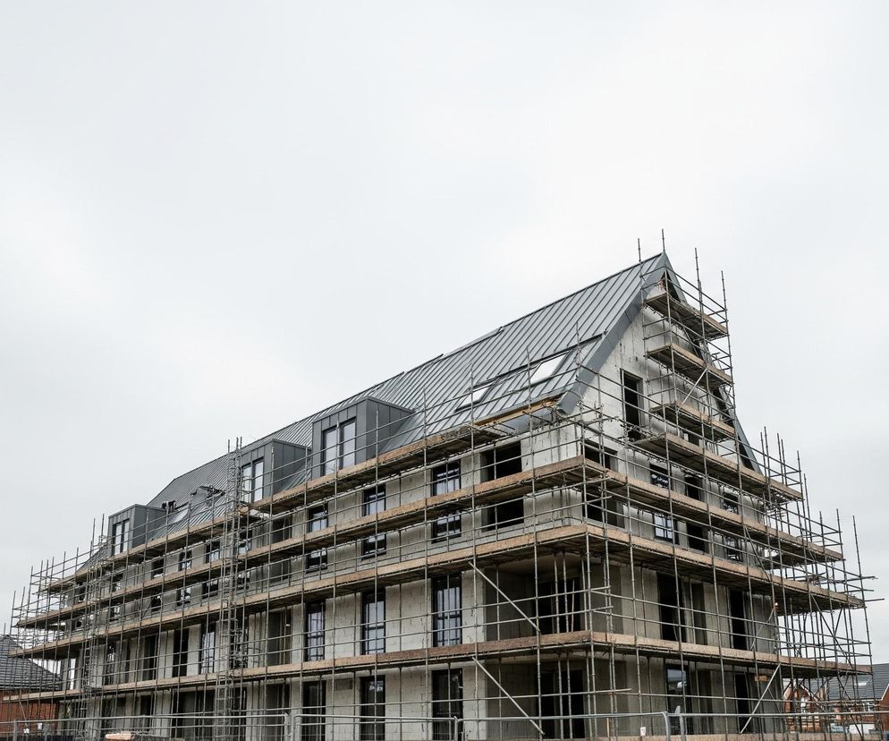 Na zdjęciu dominuje duży, wielopiętrowy budynek w trakcie budowy, pokryty rusztowaniami. Ma spadzisty, ciemnoszary dach z metalowych paneli, w którym osadzone są liczne okna dachowe i pionowe lukarny. Fasada jest w szarym kolorze, a w niej widać szereg prostokątnych, ciemnych okien.

Dookoła budynku rozciąga się metalowe rusztowanie z drewnianymi podestami, oplatające go z każdej strony. W tle, po obu stronach rozbudowywanego obiektu, widać fragmenty istniejącego już osiedla domów jednorodzinnych z ciemnymi dachami i elewacjami w odcieniach cegły oraz szarości. Niebo jest jednolicie jasnoszare, co sugeruje pochmurny dzień.