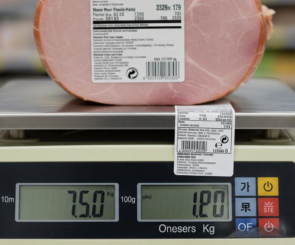 Na środku kadru znajduje się duży kawałek szynki, owinięty w folię, z białą etykietą produktową na górze, zawierającą czarny kod kreskowy i tekst. Szynka leży na metalowej, błyszczącej platformie wagi elektronicznej w kolorze kremowym. Wyświetlacz wagi pokazuje cyfry 7.50 Kg oraz 1.20, a także różne przyciski funkcyjne po prawej stronie, w tym żółty przycisk z symbolem włączania/wyłączania. Tło jest rozmyte, ale widać kolorowe półki z produktami.