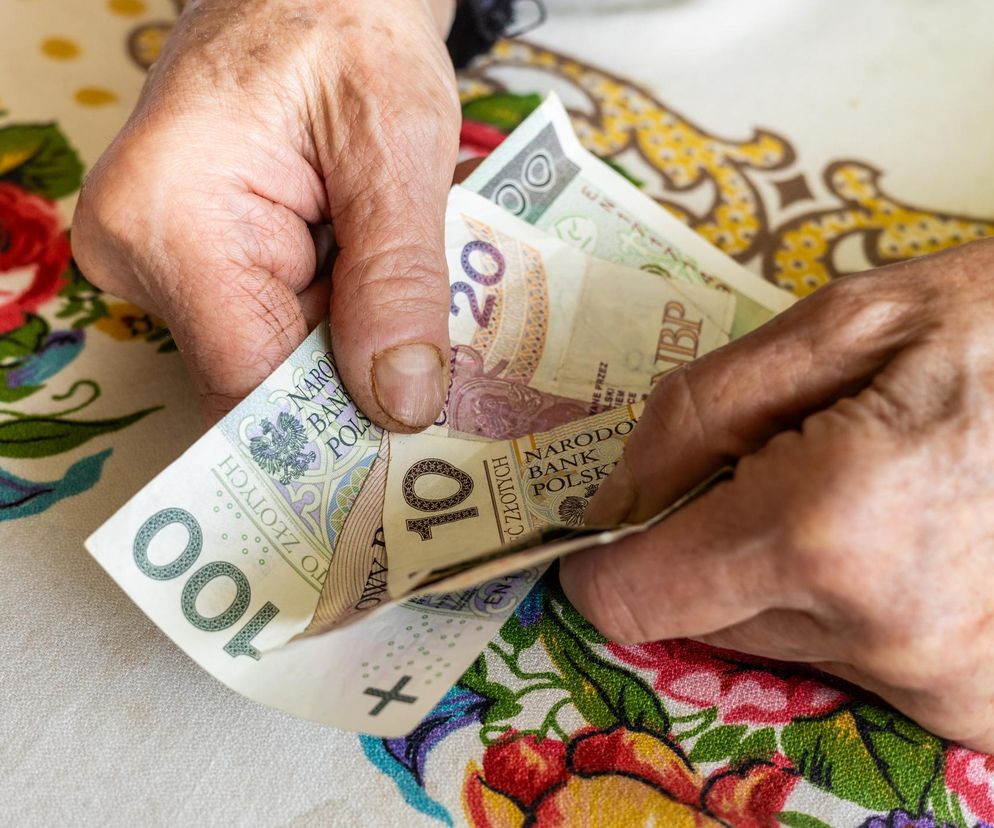 Starsze, pomarszczone dłonie osoby liczą banknoty o nominałach 100, 20 i 10 złotych, rozłożone na wzorzystym obrusie z kwiatowym motywem. Scena symbolizuje finanse osobiste i potrzebę pożyczek, o których przeczytasz więcej na Super Biznes.