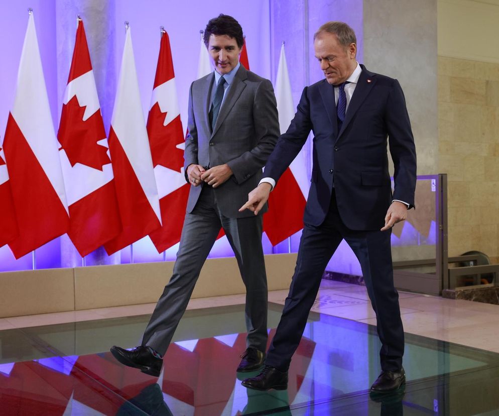 Justin Trudeau w szarym garniturze i Donald Tusk w granatowym, idący obok siebie po szklanej podłodze, z rzędem flag Kanady i Polski w tle. Tusk wskazuje palcem na dół. Temat ten jest szerzej omawiany na Super Biznes.