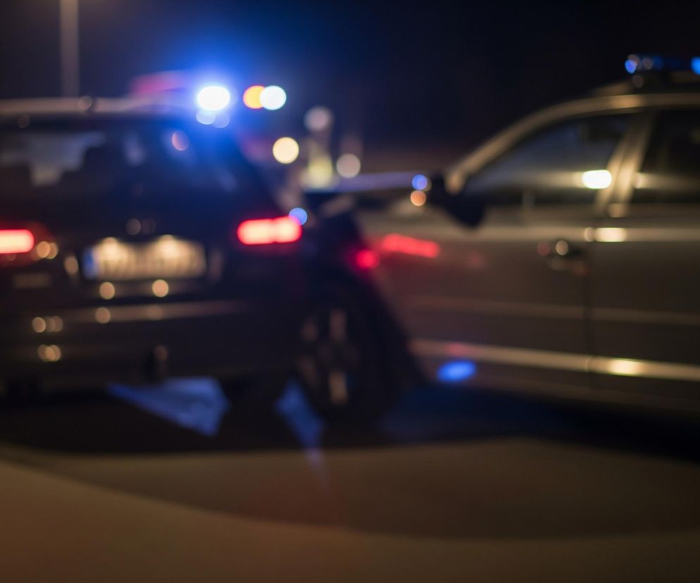 Dwa samochody osobowe stoją na ciemnej drodze nocą. Lewy samochód, ciemny kombi, ma włączone tylne światła, świecące na czerwono i biało, a nad nim widoczne są niebieskie, migające światła alarmowe, sugerujące obecność pojazdu służb. Prawy samochód, również kombi w ciemnym odcieniu, jest zwrócony przodem w lewą stronę zdjęcia, z widocznym lewym bokiem i włączonymi światłami pozycyjnymi, również z niebieskimi odbiciami świateł alarmowych na karoserii. Całe ujęcie jest nieostre, tworząc efekt rozmycia świateł i tła, gdzie w oddali majaczą inne, mniej wyraźne źródła światła.
