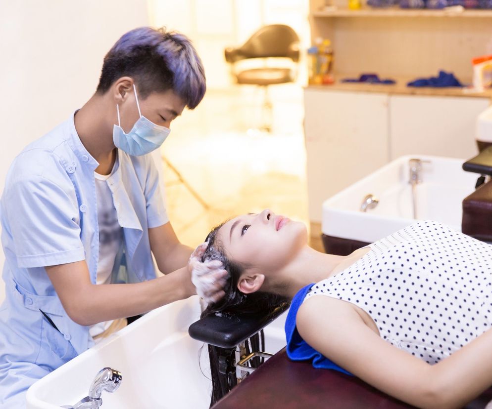 Fryzjer w maseczce ochronnej myje włosy klientce leżącej na fotelu. Grafika ilustruje ryzyko zakażeń w salonach fryzjerskich, o czym przeczytasz więcej na Poradnik Zdrowie.