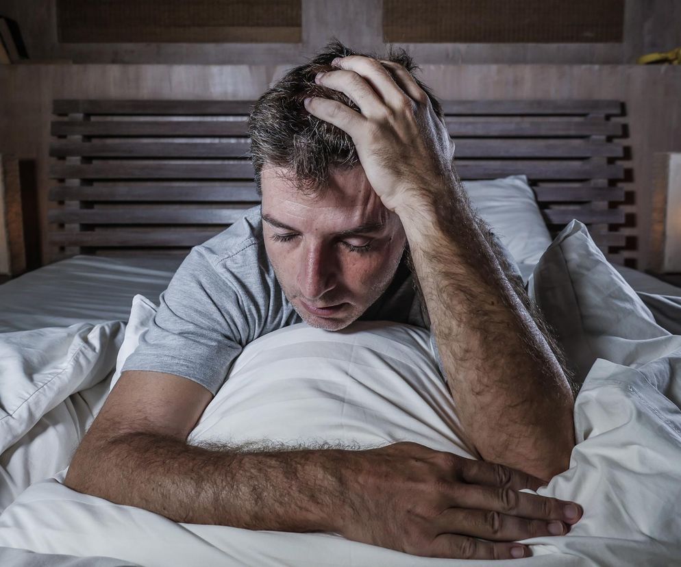 Mężczyzna z dłońmi na głowie leży w łóżku, patrząc w dół z zamyśloną, zmartwioną miną, co symbolizuje depresję lub myśli samobójcze, będące potencjalnym skutkiem ubocznym leku na łysienie, o czym informuje Poradnik Zdrowie.