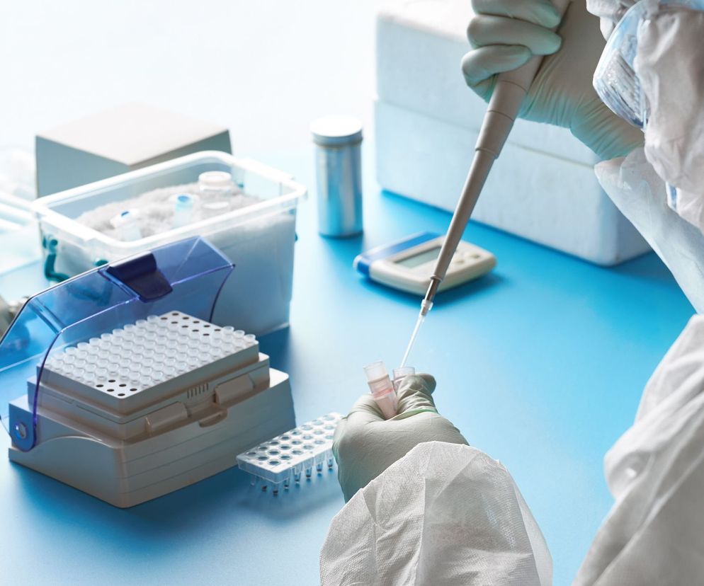 Osoba w białym kombinezonie ochronnym i rękawiczkach lateksowych pobiera pipetą ciecz z probówki, w tle widoczne laboratoryjne naczynia i pojemniki na niebieskim stole. Opis testów serologicznych i molekularnych oraz ich zastosowanie znajdziesz na portalu Poradnik Zdrowie.