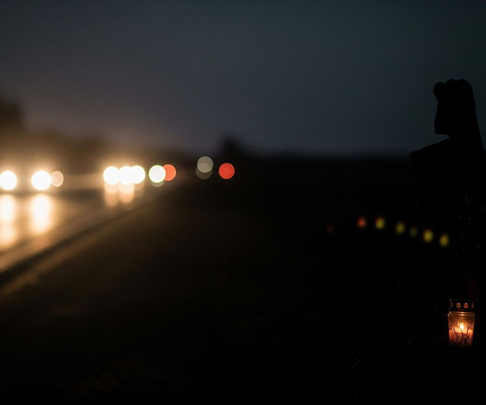 Ciemną scenę nocną z rozmywającym się tłem. Na pierwszym planie, po prawej stronie, widać wyraźnie zarys nagrobnej świecy z płonącym w środku płomieniem, rzucającym ciepłe, pomarańczowe światło. Nad świecą znajduje się ciemna sylwetka, prawdopodobnie symbolizująca postać, z wyraźnym konturem trzymanego kwiatu, prawdopodobnie lilii. W tle, po lewej stronie, rozmyte światła samochodów jadących drogą tworzą poziomy szereg jasnych, okrągłych punktów, głównie białych i pomarańczowych.