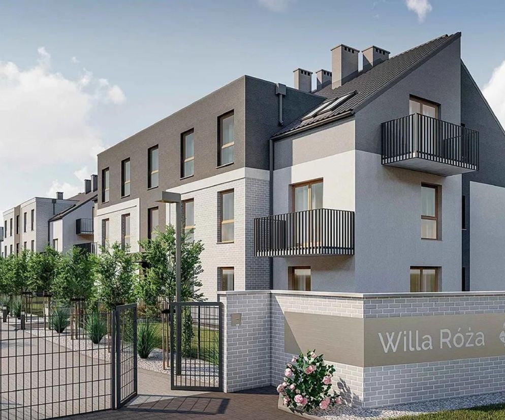 Wizualizacja nowoczesnego budynku wielorodzinnego Willa Róża z balkonami i skośnym dachem w odcieniach szarości, z dwiema osobami idącymi chodnikiem wzdłuż ogrodzenia. Na murku ceglanym widnieje nazwa obiektu, o czym przeczytasz więcej na portalu Architektura Murator Plus.