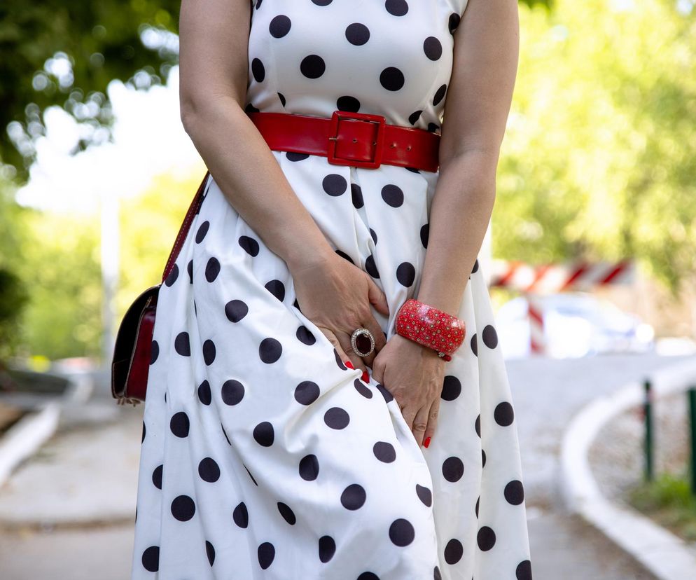 Kobieta w sukience w grochy z czerwoną torebką i paskiem, zakrywająca dłonią podbrzusze, symbolizująca problem nietrzymania moczu, o którym przeczytasz na Poradnik Zdrowie. Tło to rozmyty krajobraz miejski.