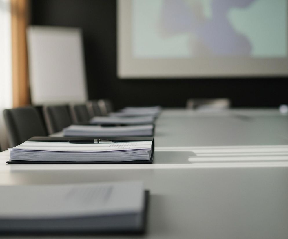 Na pierwszym planie, z lewej strony, widoczny jest szary blat stołu konferencyjnego, na którym leżą otwarte zeszyty lub notatniki w czarnych oprawach. Na jednym z nich spoczywa czarny długopis. Kolejne notatniki ustawione są w rzędzie, znikając w oddali w prawą stronę. Rząd ciemnych krzeseł z oparciami z miękkiego materiału znajduje się w tle za stołem, rozciągając się wzdłuż jego długości. W tle, po lewej stronie, widać jasne okno z białymi firankami i brązowymi zasłonami, a obok niego białą tablicę typu flipchart. Na czarnej ścianie, na wprost stołu, wyświetlany jest niewyraźny obraz w kolorze fioletowo-niebieskim, prawdopodobnie z projektora. Po prawej stronie zdjęcia, częściowo widoczna jest ciemnoniebieska flaga z żółtymi gwiazdami.