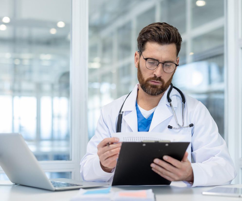 Mężczyzna w białym fartuchu lekarskim i okularach, z brodą, skupiony na dokumentach na podkładce, trzymając je w dłoniach, obok laptop. Zdjęcie ilustruje temat utraty prawa do darmowego leczenia z NFZ, o czym można przeczytać na portalu Poradnik Zdrowie.