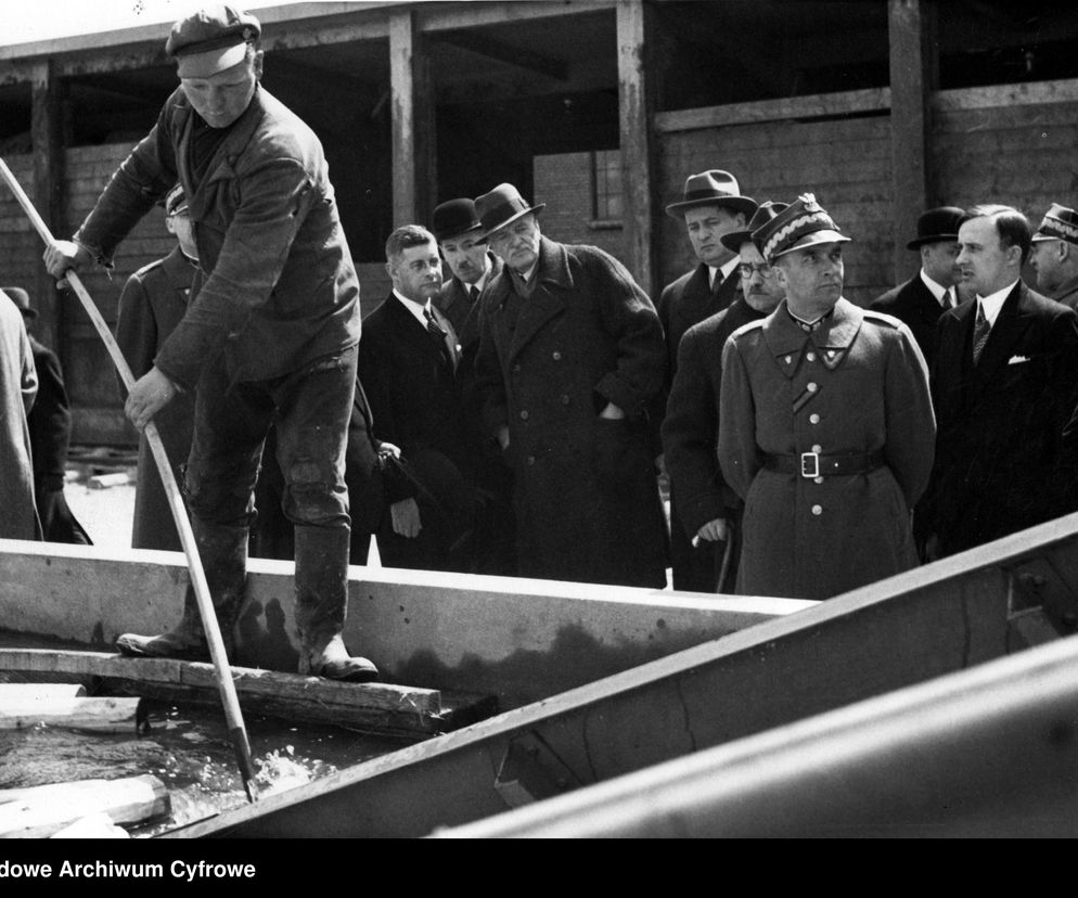 Urzędnicy i żołnierz w mundurze obserwują pracownika budowy, który w brudnym ubraniu i czapce pracuje na konstrukcji Centralnego Okręgu Przemysłowego. Historyczny moment modernizacji polskiej gospodarki, o którym więcej można przeczytać na Super Biznes.