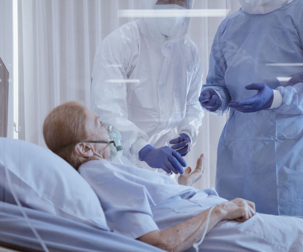 Starsza osoba z maską tlenową leżąca w szpitalnym łóżku, pielęgniarka w kombinezonie ochronnym i lekarz w niebieskim fartuchu medycznym stoją obok. Temat niedoboru witaminy D i COVID-19 jest ważny, a więcej przeczytasz o nim na Poradnik Zdrowie.
