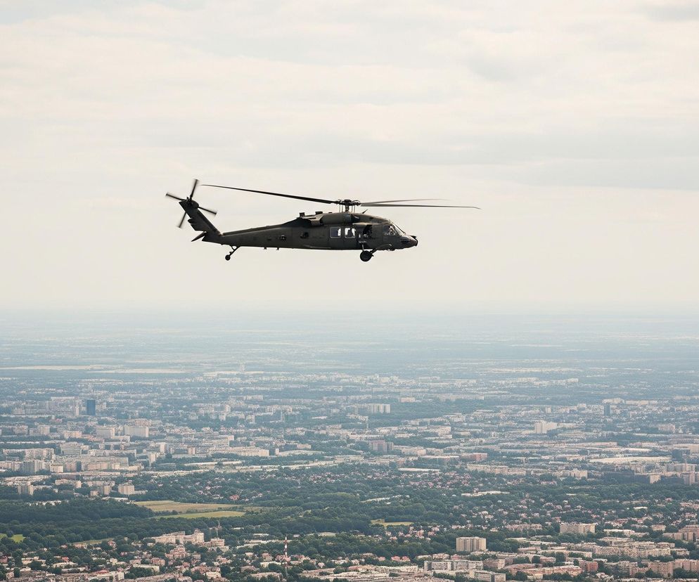 W centralnej części ujęcia znajduje się ciemnozielony helikopter z głównym i tylnym wirnikiem. Maszyna jest w locie, widoczna z boku, lekko odchylona w lewo. Pod nią rozciąga się rozległy, zamglony krajobraz miejski, z licznymi budynkami i zielonymi obszarami drzew. Górna część obrazu to jasne, zamglone niebo z nielicznymi, rozmyty obłokami.