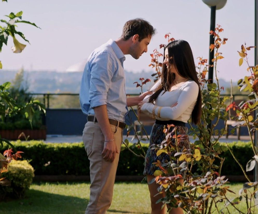 Młody mężczyzna w jasnoniebieskiej koszuli i beżowych spodniach rozmawia z kobietą o długich, ciemnych włosach, ubraną w białą bluzkę i kolorową spódnicę. Stoją blisko siebie w ogrodzie, otoczeni zielenią i krzewami. Scena ilustruje dramatyczne wydarzenia z serialu Miłość i nadzieja, gdzie Bahar stawia ultimatum Leventowi. Więcej szczegółów na Super Seriale.