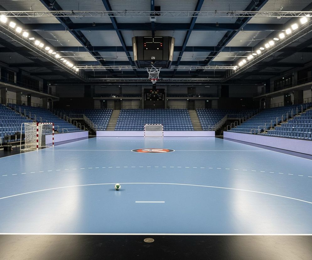 Duża, pusta hala sportowa z jasnoniebieskim parkietem zajmuje centrum kadru. Na parkiecie widoczne są białe linie boiska, a na środku, bliżej pierwszego planu, leży piłka nożna w zielono-białe łaty. Po obu stronach boiska rozciągają się rzędy pustych, granatowych krzeseł, ułożonych amfiteatralnie. Na dalszym planie znajdują się dwie bramki sportowe z biało-czerwonymi słupkami i siatkami. Powyżej trybun, pod sufitem, zawieszony jest elektroniczny zegar wyników, a cała hala jest oświetlona rzędami jasnych lamp umieszczonych na metalowych konstrukcjach sufitu.
