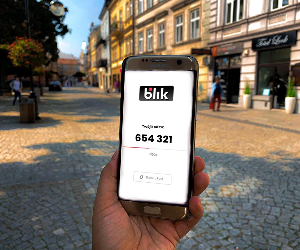 Dłoń trzyma smartfon Samsung z wyświetlonym kodem BLIK 654 321 na tle brukowanej ulicy z kamienicami. Scena symbolizuje problemy z płatnościami BLIK po ataku DDoS, o których można przeczytać na Super Biznes.