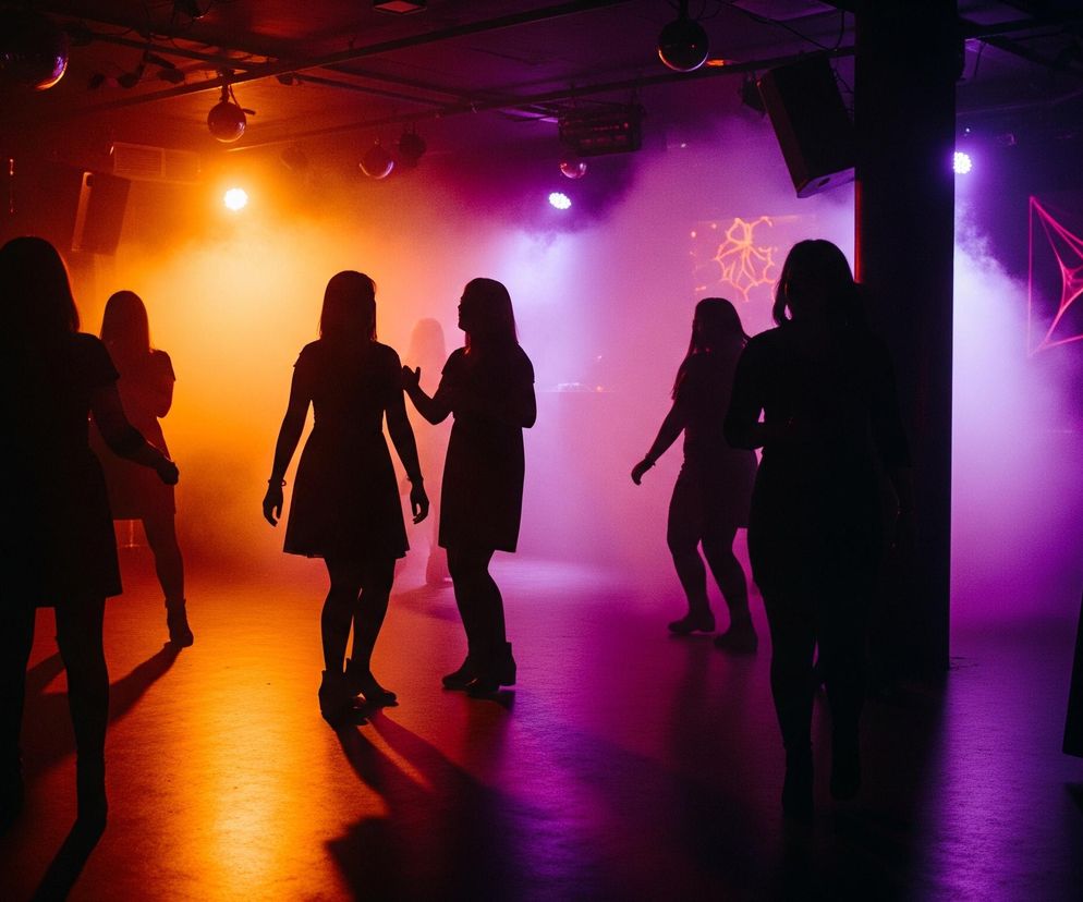 Grupę ludzi, głównie kobiet, tańczących w słabo oświetlonym pomieszczeniu, prawdopodobnie klubie lub na imprezie. Sylwetki osób są ciemne, niemal czarne, oświetlone od tyłu intensywnym, kolorowym światłem, które tworzy pomarańczowo-fioletowe odcienie dymu wypełniającego przestrzeń. Po prawej stronie, na tle fioletowego światła, widać dekorację przypominającą pajęczynę z pająkiem oraz maskę w kształcie kota z pomarańczowymi elementami, leżącą na czarnym stoliku. Na suficie widoczne są kule dyskotekowe i reflektory, a na ścianie po lewej stronie znajduje się ekran z jasnym, niewyraźnym napisem.