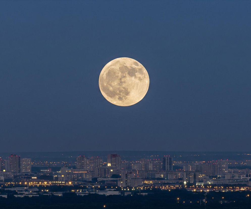 Pełnia księżyca dominująco zajmuje centralną część nieba, przedstawioną w odcieniu głębokiego granatu. Powierzchnia księżyca jest szczegółowo widoczna, z licznymi kraterami i nierównościami, które nadają jej chropowaty, trójwymiarowy wygląd. Poniżej rozpościera się panorama miejska z licznymi wieżowcami i budynkami, z których większość ma zapalone światła. Horyzont miasta jest usiany drobnymi, świecącymi punktami, a na pierwszym planie widoczne są ciemne sylwetki drzew i zieleni.