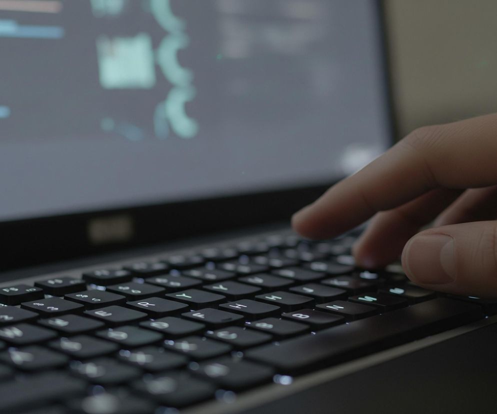 Dłoń z palcami opartymi na czarnej klawiaturze laptopa jest głównym elementem obrazu, widoczna w prawym dolnym rogu. Ekran laptopa, znajdujący się w tle, wyświetla jasne linie i kształty przypominające kod lub dane, rozmyte i niewyraźne, w odcieniach szarości i błękitu. Klawisze mają białe podświetlenie liter i symboli, a niektóre z nich są wyraźnie widoczne, jak np. F9, F10, R, T, Y, H, B, N, M. Tło za ekranem jest nieostre, widoczne są na nim zielone punkty światła.