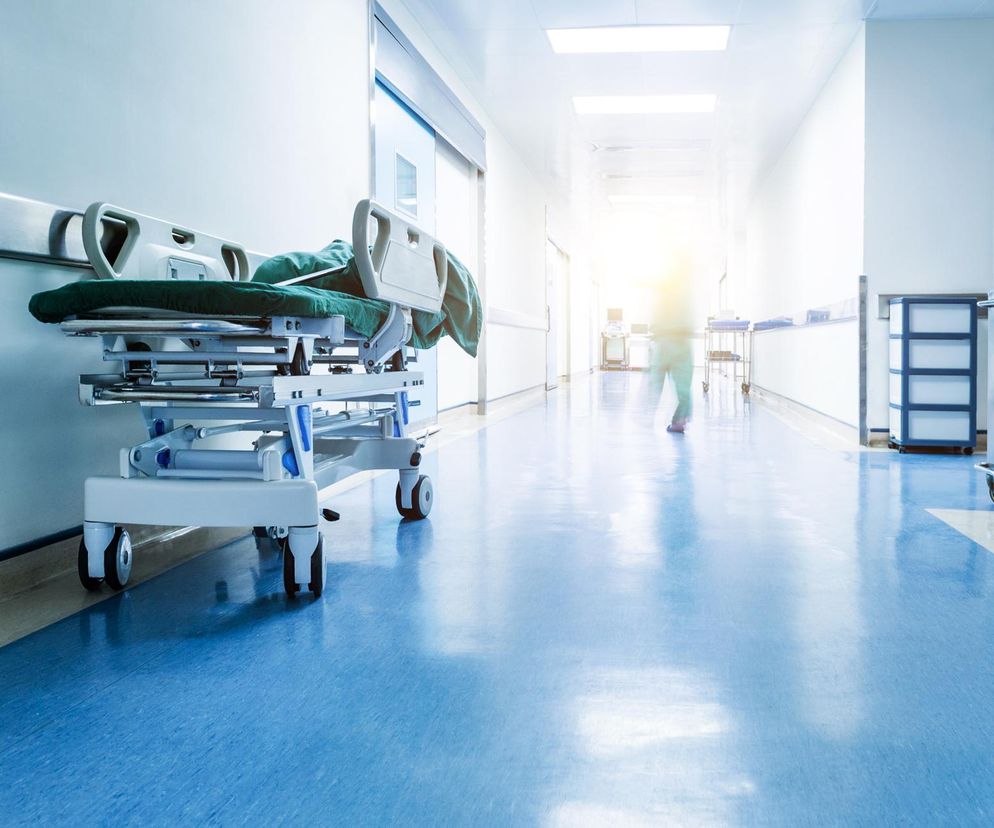 Jasny, sterylnie wyglądający korytarz szpitalny z pustym łóżkiem medycznym na kółkach po lewej stronie i rozmytą postacią w tle, symbolizujący problemy finansowe służby zdrowia i wstrzymywanie przyjęć pacjentów, o których można przeczytać na Poradnik Zdrowie.