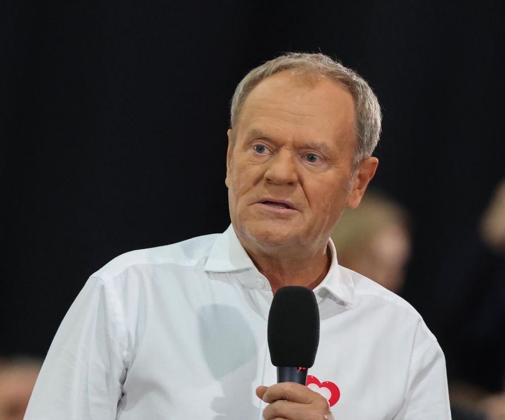 Donald Tusk, premier, w białej koszuli, z mikrofonem w dłoni, podczas konferencji prasowej w Piotrkowie Trybunalskim, dyskutuje o mieszkaniach dla ministrów i ich wysokich czynszach. O szczegółach przeczytasz na Super Biznes.
