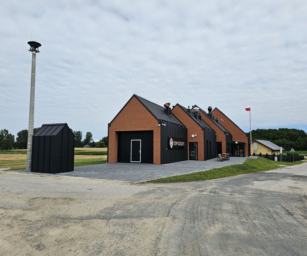Nowoczesna remiza OSP Kozjaty z cegły i czarnej blachy, z wyraźnym logo jednostki, osadzona w wiejskim krajobrazie, otoczona polami. Ten funkcjonalny budynek w województwie kujawsko-pomorskim pokazuje kierunki rozwoju architektury publicznej, o czym więcej przeczytasz na portalu Architektura Murator Plus.