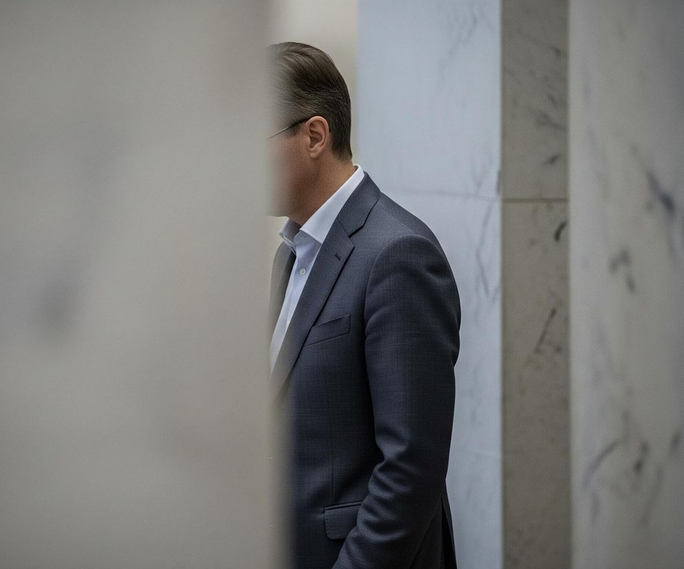 Mężczyzna w ciemnym garniturze i jasnej koszuli jest częściowo widoczny po prawej stronie kadru, z jego twarzą zasłoniętą przez jasny, niewyraźny element na pierwszym planie. Ubrany jest w szarą marynarkę z klapami i białą koszulę z rozpiętym guzikiem pod szyją, a także nosi okulary. Tło stanowią marmurowe ściany w odcieniach bieli i szarości, z widocznymi ciemniejszymi żyłkami, które nadają im teksturę.