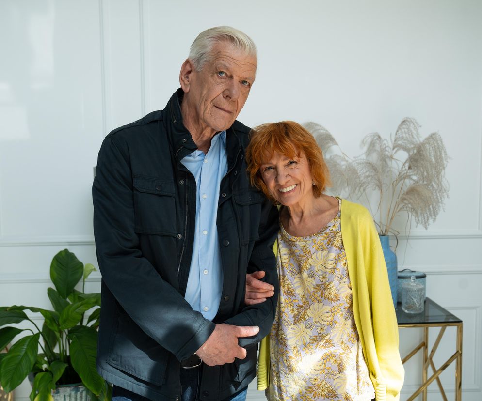 Ewa Kolasińska jako pani Ola i Marcin Sosnowski jako pan Mirek w scenie z serialu M jak miłość. Para aktorów uśmiecha się do obiektywu, a aktorka, ubrana w kwiecistą bluzkę i żółty sweter, opiera głowę na ramieniu aktora. Więcej o ich losach na Super Seriale.