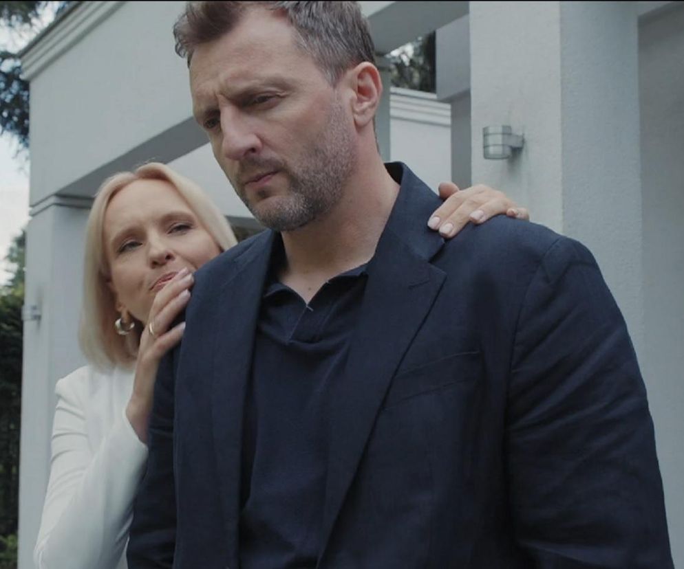 Bartek (Arkadiusz Smoleński) z zamyślonym wyrazem twarzy, Dorota (Iwona Rejzner) opiera dłoń na jego ramieniu, patrząc z troską. Aktorzy z M jak miłość stoją przed jasnym budynkiem, symbolizującym ich małżeńskie rozterki i plany biznesowe. Więcej o ich losach na Super Seriale.