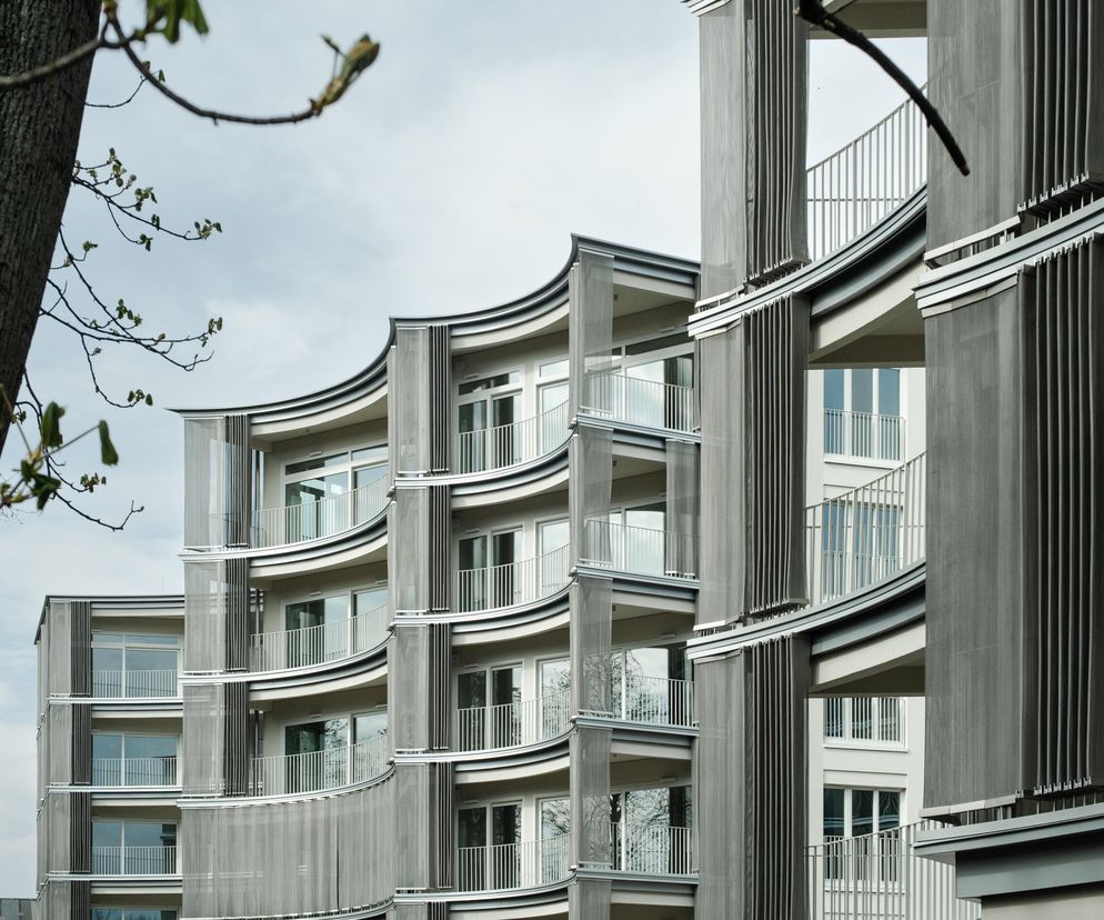 Zbliżenie na falujące, półkoliste balkony budynków Perfumiarni w Poznaniu, z metalowymi żaluzjami, częściowo przesłonięte pniem i gałęziami drzewa. To nominowana do Życia w Architekturze 2025 realizacja JEMS Architekci, którą opisuje Architektura Murator Plus.