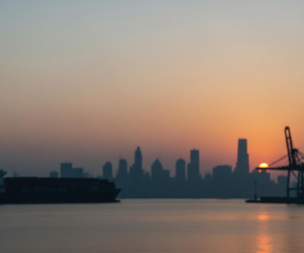 Na pierwszym planie, w dolnej części kadru, rozciąga się szeroka tafla wody o lekko falującej powierzchni. Po lewej stronie, na wodzie, unosi się ciemny statek kontenerowy, którego sylwetka jest niewyraźna. Horyzont dominuje miasto o zarysie wieżowców, a za nim, po prawej stronie, wschodzi słońce w kolorze pomarańczowo-żółtym. Po prawej stronie, na tle słońca, znajdują się ciemne sylwetki portowych dźwigów, a niebo nad horyzontem przechodzi od pomarańczowych i różowych odcieni w dolnej części do jaśniejszych błękitów u góry.