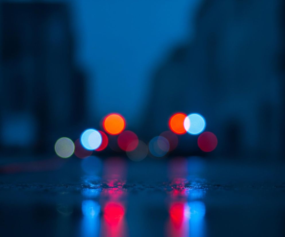 W centralnej części obrazu widoczne są rozmyte, okrągłe światła o barwach czerwonej, pomarańczowej, niebieskiej i jasnozielonej, tworzące efekt bokeh. Światła te odbijają się od mokrej, ciemnej powierzchni, tworząc jaśniejsze, pionowe refleksy w dolnej części kadru. Tło, głęboko niebieskie, ukazuje rozmyte, ciemne kształty, sugerujące sylwetki budynków.