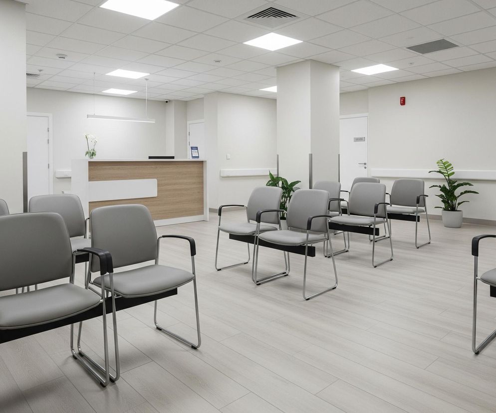 Jasne, nowoczesne pomieszczenie z rzędami szarych krzeseł, ustawionymi na panelowej podłodze w odcieniach jasnego drewna, rozciąga się w głąb. Po lewej stronie widać ścianę, a po prawej, na końcu pomieszczenia, znajduje się recepcyjna lada z drewnianym frontem i jasnym blatem, za którą stoi wazon z białymi kwiatami. Sufit jest kasetonowy, z równomiernie rozmieszczonymi, prostokątnymi oprawami oświetleniowymi emitującymi jasne światło. Przy ścianach widoczne są białe listwy ochronne oraz zielone rośliny w doniczkach, dodające akcentów kolorystycznych do stonowanego wnętrza.