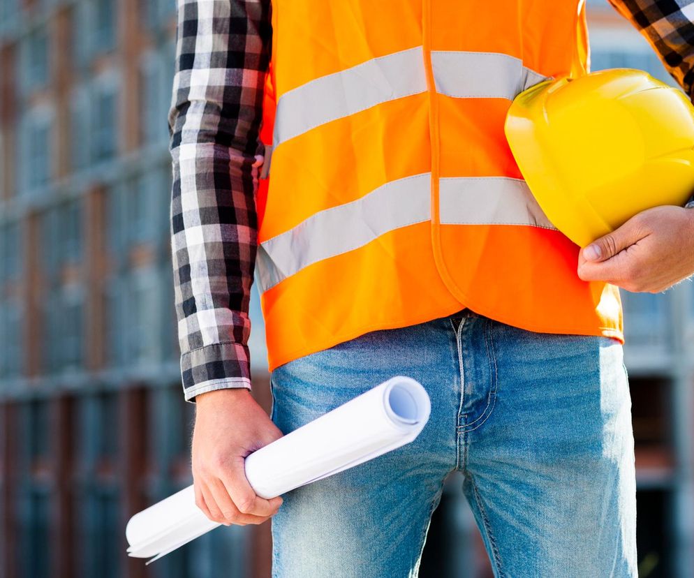 Pracownik budowlany w pomarańczowej kamizelce odblaskowej, z żółtym kaskiem w ręku i zrolowanymi planami, na tle placu budowy. Artykuł na Super Biznes informuje o prawach pracownika do rekompensaty za chorobę zawodową.