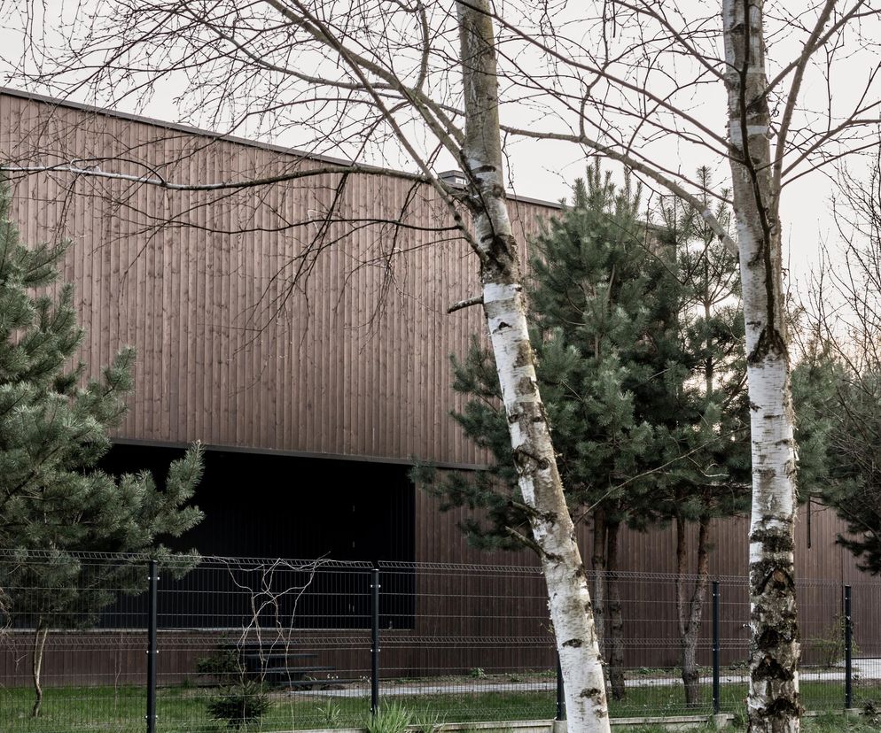 Bryła ZBUCKA HOUSE w Karakulach, pokryta drewnianą elewacją w odcieniu pni sosen, wpisująca się w leśny krajobraz. Czarne, cofnięte fragmenty elewacji akcentują funkcje, a całość ukryta jest za zielenią i siatkowym ogrodzeniem. O projekcie przeczytasz na Architektura Murator Plus.