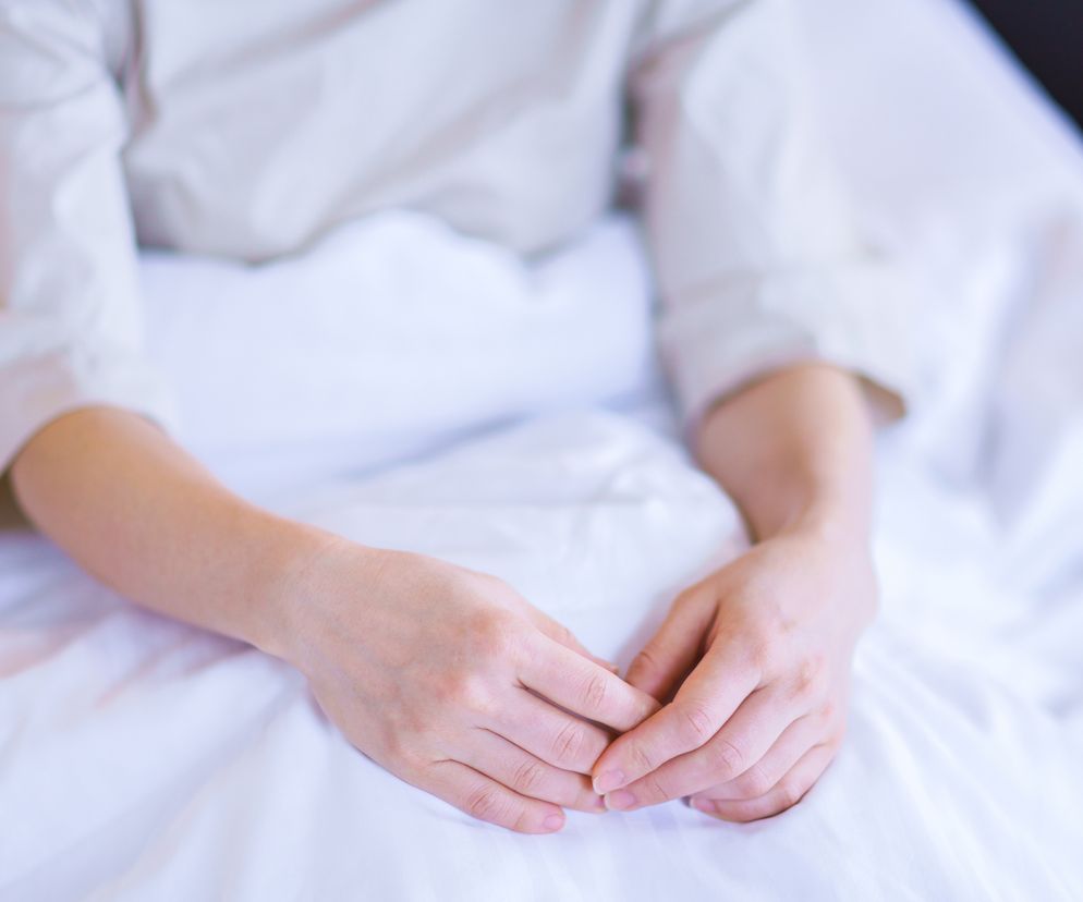 Kobieta w średnim wieku siedzi owiniętą białą pościelą na łóżku.