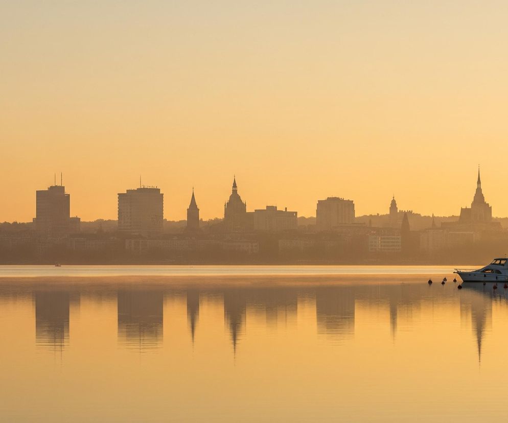 Horyzont dzieli obraz na dwie części: górną, przedstawiającą rozległe niebo, oraz dolną, ukazującą spokojną taflę wody z odbiciami. W tle, na całej szerokości horyzontu, widoczna jest sylwetka miasta z licznymi wysokimi budynkami i wieżami, w tym kilkoma z ostrymi, spiczastymi dachami. Odcienie pomarańczu i złota dominują całą scenę, tworząc wrażenie wczesnego poranka lub późnego wieczoru, z rozproszonym światłem, które rozjaśnia mglistą atmosferę.

Na pierwszym planie po prawej stronie, przy brzegu, cumują liczne jachty i łodzie motorowe, z których niektóre mają maszty sięgające niemal do nieba, a ich odbicia są wyraźnie widoczne na wodzie. Po lewej stronie, na tafli wody, widoczny jest pojedynczy niewielki pływający obiekt, prawdopodobnie boja, a dalej kilka innych, mniejszych elementów. Całość odbija się w lustrzanej powierzchni wody, powtarzając kształty budynków i łodzi w lekko rozmytej formie, z drobnymi falami tworzącymi poziome linie na odbiciach.