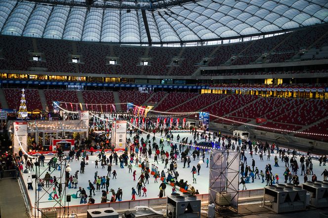 Płyta PGE Narodowego zamieniona w Zimowe Miasteczko z dwoma lodowiskami, na których ślizgają się liczne rodziny i dzieci. Widoczna jest ozdobiona choinka oraz iluminacje świetlne na trybunach stadionu. Na Poradnik Zdrowie znajdziesz więcej informacji o atrakcjach aktywnego wypoczynku.