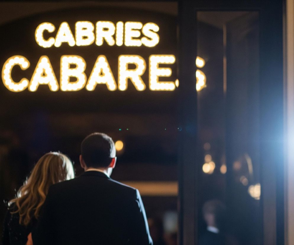 W centrum obrazu widoczne są plecy dwóch osób – mężczyzny i kobiety, idących w kierunku przeszklonych drzwi, za którymi rozciąga się ciemne wnętrze. Za drzwiami, w głębi, świeci duży neon z białymi, podświetlanymi żarówkami, tworzącymi napisy: CABRIES w górnej linii i CABARE w dolnej, z brakującą ostatnią literą. Prawa strona obrazu jest silnie rozjaśniona przez jasne, białe światło, które tworzy refleksy i niebieskie poświaty.