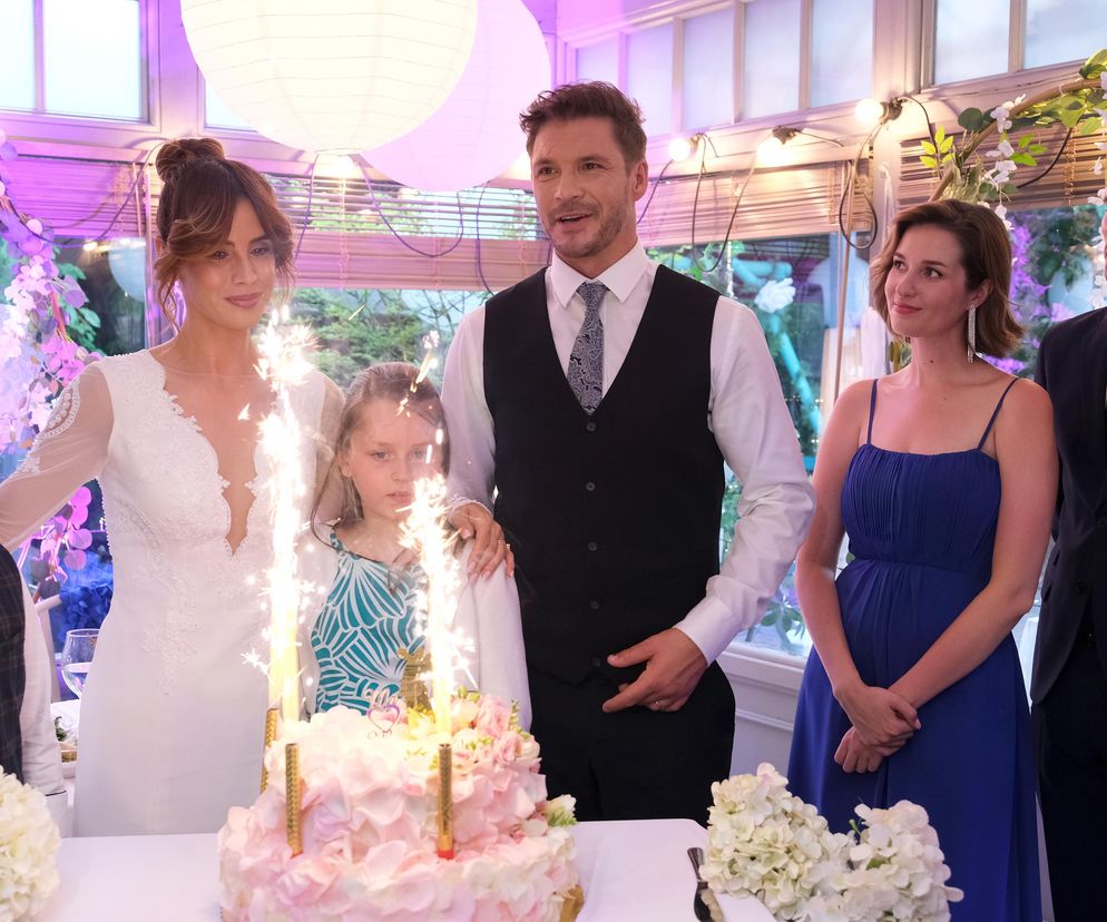 Uśmiechnięta Michalina Sosna jako Kama w białej sukni ślubnej i Marcin Roznerski w kamizelce i białej koszuli, stojący przy torcie weselnym z fontannami. Obok nich dzieci Laura Jankowska i Szymek, a także Alina Szczegielniak w niebieskiej sukience oraz Maurycy Popiel w garniturze. Zobacz, co wydarzy się na weselu Kamy i Marcina na Super Seriale.