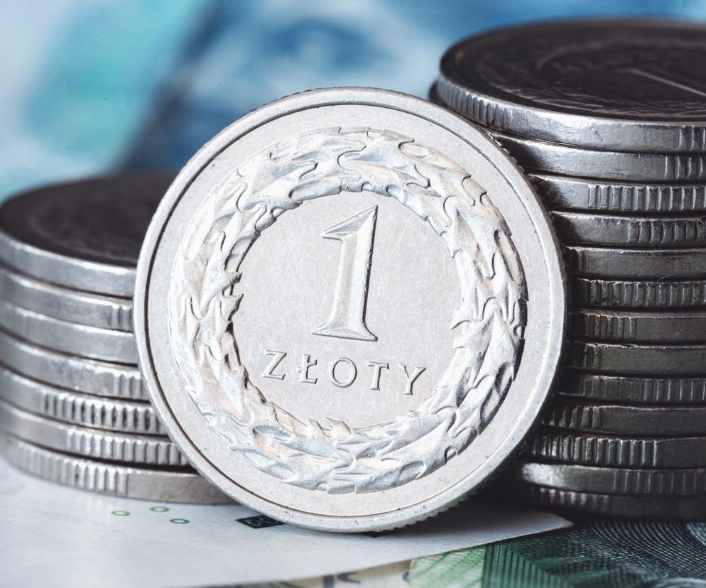 Moneta 1 złoty na tle stosu innych monet i banknotów symbolizująca finanse, oszczędności oraz kwestie socjalne. Obraz ilustruje kontekst artykułu o świadczeniach socjalnych jak 500 plus czy propozycję 500 zł dla pracujących, o czym można przeczytać na Super Biznes.