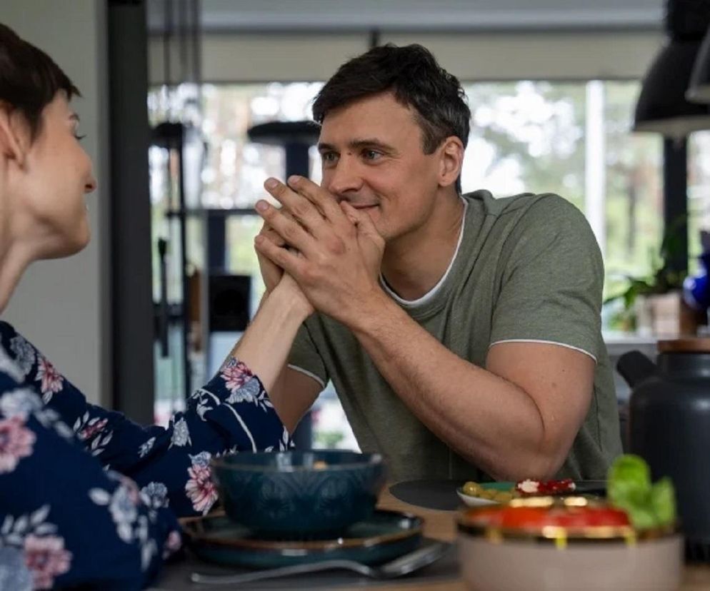 Krzysztof Radwan (Mateusz Damięcki) i Blanka (Pola Gonciarz) siedzą przy stole, trzymając się za ręce. Mężczyzna uśmiecha się, patrząc na kobietę w kwiatowej koszuli nocnej. Romantyczny nastrój wieczoru zakochanych, o którym można przeczytać na Super Seriale.