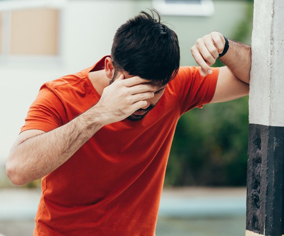Mężczyzna w pomarańczowej koszulce, z ręką na czole, opierający się o słupek, symbolizuje problemy z zawrotami głowy i pamięcią, które mogą ostrzegać przed rakiem jądra. Więcej o objawach i diagnostyce przeczytasz na Poradniku Zdrowie.