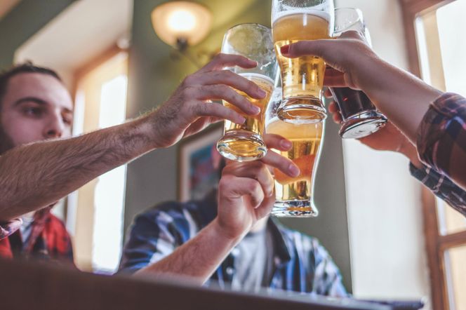 Grupa przyjaciół wznosi toast piwem w szklankach, celebrując wspólną chwilę. Dłoń z pomalowanymi paznokciami unosi ciemne piwo, podczas gdy pozostałe dłonie trzymają jasne piwo. Kontekst dotyczy wpływu nawet umiarkowanego spożycia alkoholu na zdrowie, o czym można przeczytać na portalu Poradnik Zdrowie.