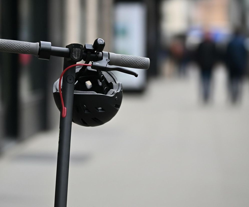 Przednia część czarnej hulajnogi elektrycznej dominuje w lewej części kadru. Na kierownicy, z szarymi, perforowanymi uchwytami, zawieszony jest czarny kask z otworami wentylacyjnymi, a czerwony kabel biegnie w dół wzdłuż kierownicy. W tle, w silnym rozmyciu, widać ulicę z niewyraźnymi sylwetkami ludzi i zarysami budynków.