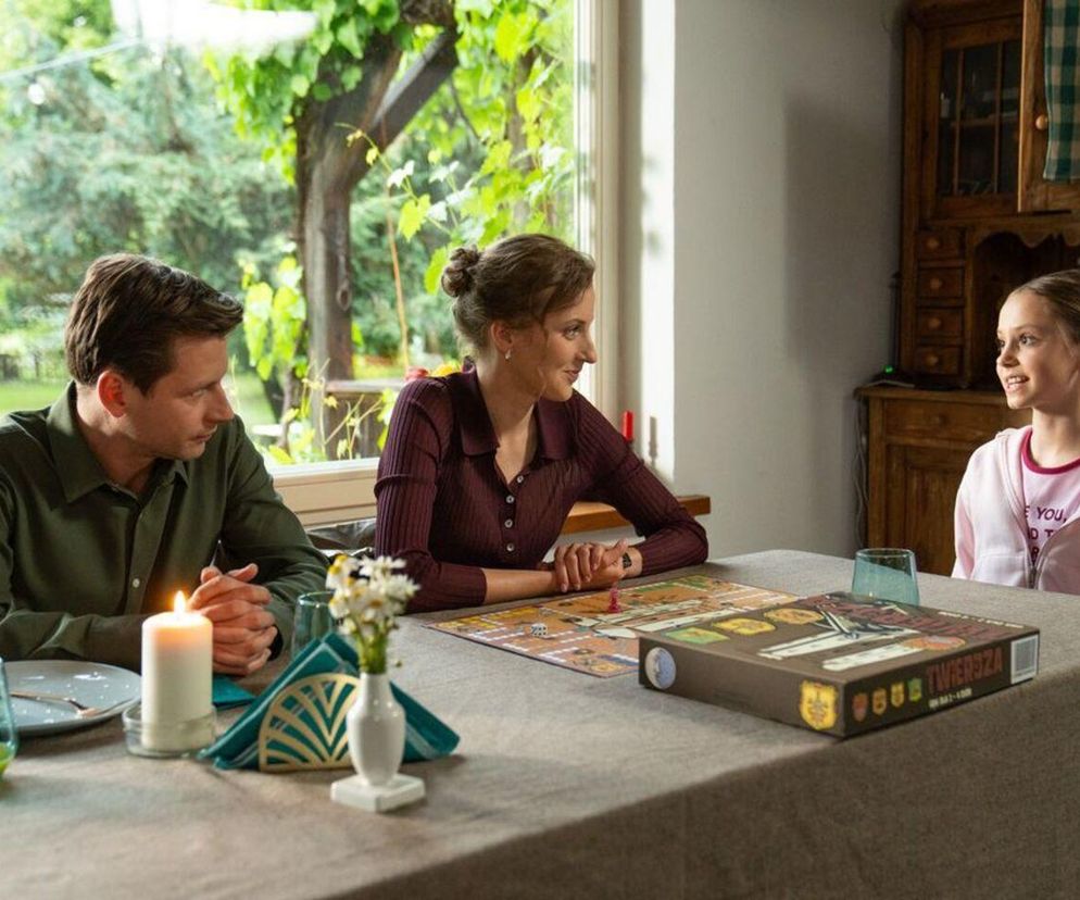 Adam Karski, Natalka Mostowiak i Hania w radosnej scenie przy stole z grą planszową, symbolizującej rodzinne szczęście i nowe początki. Hania, uśmiechnięta, prawdopodobnie doprowadziła do ujawnienia romansu Natalki i Adama, co widać na Super Seriale.